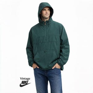 May include: A dark green hooded anorak jacket with a front pocket and a small Nike logo. The jacket has a half-zip closure and is worn with blue jeans. The word "Vintage" and the Nike logo are visible.
