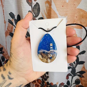 Puede incluir: Un collar con colgante en forma de lágrima con una escena de playa en miniatura. El colgante de resina tiene un fondo azul océano con un faro, conchas marinas y arena. El collar tiene un cordón negro.