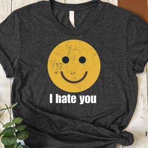 May include: Dark gray t-shirt featuring a distressed yellow smiley face with black eyes and a curved mouth. Below the smiley face, the words "I hate you" are printed in white. The shirt has short sleeves and a crew neck.