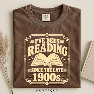 May include: A brown short-sleeve t-shirt with a cream-colored graphic print featuring an open book icon and the text "I'VE BEEN READING SINCE THE LATE 1900s." The shirt has a crew neckline and a Comfort Colors brand label at the inner collar.