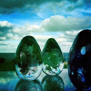 May include: Three glass egg-shaped sculptures with internal abstract designs. The two front sculptures are green, while the third is dark blue. The background features a blue sky with white clouds and a body of water.