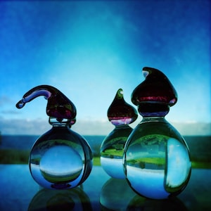 Könnte beinhalten: Drei klare Glasflaschen mit dunkelroten und violetten Stöpseln. Die Flaschen haben einen runden Boden und reflektieren den Himmel und die Landschaft. Der Hintergrund ist ein hellblauer Himmel und ein Gewässer.