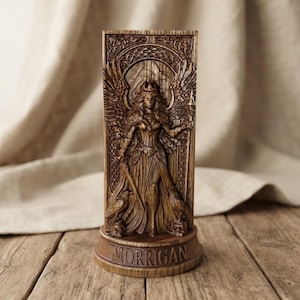 May include: A rectangular brown wooden plaque featuring a relief carving of a winged figure holding a spear and sword, flanked by two ravens and skulls. The base of the plaque is inscribed with the text MORRIGAN in raised capital letters.