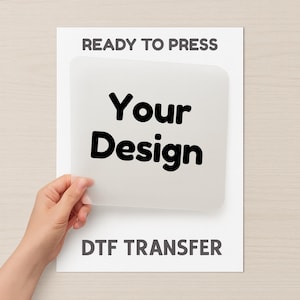 Könnte beinhalten: Ein weißes Blatt mit dem Text "READY TO PRESS" oben und "DTF TRANSFER" unten. Die Worte "Your Design" stehen in der Mitte auf einem hellfarbigen Quadrat. Eine Hand hält das Blatt.