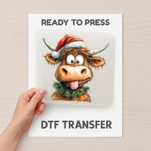 May include: A DTF transfer sheet with a cartoon Highland cow wearing a Santa hat and a green garland. The cow has a pink tongue sticking out. The text "READY TO PRESS" and "DTF TRANSFER" are printed on the sheet.