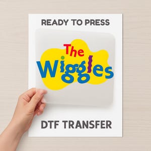 May include: A white sheet with the text "READY TO PRESS" at the top and "DTF TRANSFER" at the bottom. The centre features a yellow blob with the words "The Wiggles" in blue, red, and purple.
