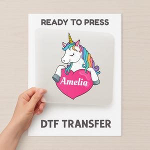 Könnte beinhalten: Ein DTF-Transferbogen mit einem Cartoon-Einhorn, Regenbogenmähne und -schwanz, das ein rosa Herz mit dem Namen "Amelia" hält. Der Text "READY TO PRESS" und "DTF TRANSFER" ist auf weißem Hintergrund gedruckt.