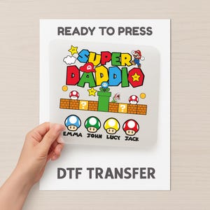 Könnte beinhalten: Ein DTF-Transfer mit einem Super Mario-Themen-Design, einschließlich Mario, Pilzen, Sternen und dem Text "SUPER DADDIO". Die Namen Emma, John, Lucy und Jack sind ebenfalls vorhanden. Die Wörter "READY TO PRESS" und "DTF TRANSFER" sind ebenfalls aufgedruckt.