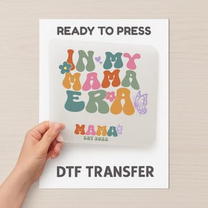 May include: A DTF transfer sheet with the text "IN MY MAMA ERA" in colourful, retro-style lettering, along with the word "MAMA EST 2025". The words "READY TO PRESS" and "DTF TRANSFER" are also printed on the white background.