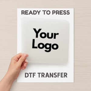 May include: A white DTF transfer sheet with the text "READY TO PRESS" at the top and "DTF TRANSFER" at the bottom. The centre features a transparent area with the words "Your Logo" in bold black font. A hand is holding the transparent area.
