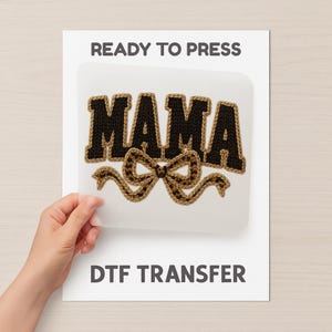 May include: A DTF transfer sheet featuring the word "MAMA" in black, textured letters with a brown border. A brown bow with a leopard print design is below the word. The words "READY TO PRESS" and "DTF TRANSFER" are printed on the white background.