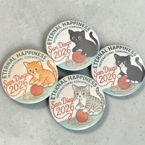 May include: Four round buttons with cat illustrations and the text "Eternal Happiness International Convention San Diego 2026". Each button features a different cat design, playing with a ball of yarn. The buttons have a light blue and orange color scheme.