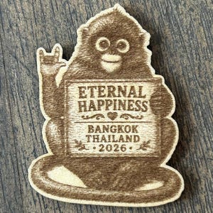 May include: A wooden souvenir featuring a cartoon monkey holding a sign. The sign reads "ETERNAL HAPPINESS BANGKOK THAILAND 2026." The monkey is brown with a smiling face and a hand gesture. The background is a wooden surface.