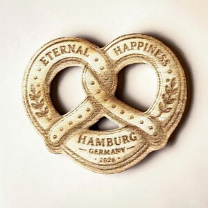 May include: A wooden pretzel-shaped ornament with the words "ETERNAL HAPPINESS" and "HAMBURG GERMANY" engraved on it. The ornament has a light brown color and includes the year "2024".