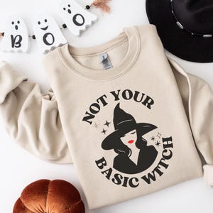 Puede incluir: Sudadera beige con la frase "NOT YOUR BASIC WITCH" y un gráfico de una bruja. La bruja lleva un sombrero negro, pintalabios rojo y tiene el pelo negro. Decoraciones de Halloween en el fondo, incluido un sombrero negro.