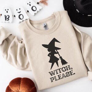 Puede incluir: Sudadera beige con un gráfico de bruja negra sosteniendo una escoba y el texto "WITCH, PLEASE." La sudadera es parte de un tema de Halloween, con un sombrero negro y decoraciones de fantasmas.