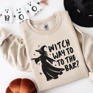 May include: Beige sweatshirt with the text "WITCH WAY TO THE BAR?" and a black witch silhouette. The sweatshirt is part of a Halloween-themed collection. A black hat and decorative ghosts are in the background.