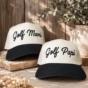 Può includere: Due cappellini da baseball con corona color bianco sporco e visiera nera. Un cappellino presenta la scritta ricamata in nero Golf Mami, mentre l'altro presenta la scritta ricamata in nero Golf Papi. Entrambi i cappellini hanno una visiera curva.