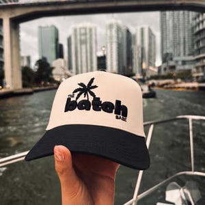 May include: A cream-colored trucker hat with a black brim and black embroidery. The front panel features a black palm tree icon above the text THE BATCH BABE in a bold, rounded font. The hat has a structured front panel and a solid black visor.
