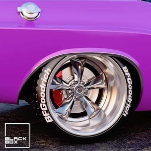 Puede incluir: Primer plano de una rueda de coche clásico. La llanta cromada rodea una pinza de freno roja. El neumático negro lleva la inscripción "BFGoodrich" en blanco. La carrocería del coche es morada. Incluye el logo "BLACK BOX".