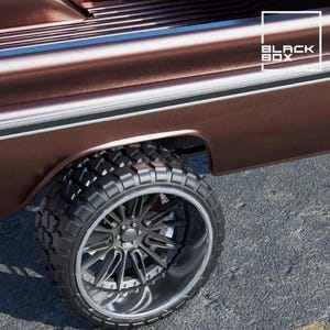 May include: A close-up of a brown truck's wheel and body. The large black tyre has a rugged tread, and the rim is silver. The truck's body has a metallic finish with a white stripe. The text "BLACK BOX" is in the upper right corner.