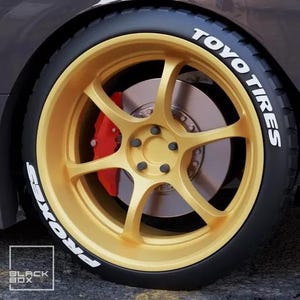 May include: Close-up of a car wheel with a gold-colored rim and a black tire. The tire has white text that reads "TOYO TIRES" and "PROXES". A red brake caliper is visible behind the spokes.