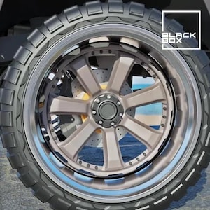 May include: Close-up of a vehicle wheel with a black tire and a silver and bronze-colored rim. The rim has a multi-spoke design and a central hub. The tire has a rugged tread pattern. The text "BLACK BOX" is in the upper right corner.