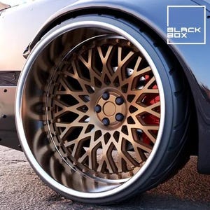 May include: Close-up of a car wheel with a bronze-colored, intricate spoke design. The wheel has a polished silver rim and a black tire. The brake calipers are red. The text "BLACK BOX" is in a white box in the upper right corner.