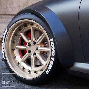 May include: Close-up of a car wheel with a gold-coloured rim and black tyre. The tyre has white lettering that reads "TOYO TIRES." The brake calipers are red. The car's body is dark grey.