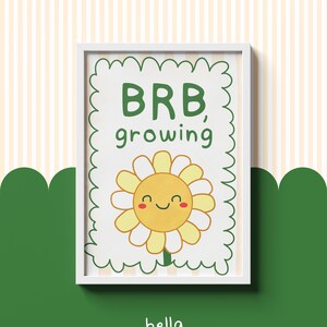 Könnte beinhalten: Ein gerahmter Druck mit einer lächelnden Cartoon-Sonnenblume mit gelben Blütenblättern und einem grünen Stiel. Der Text "BRB, growing" steht über der Blume. Der Druck befindet sich in einem weißen Rahmen, mit den Worten "bella & bill" unten.