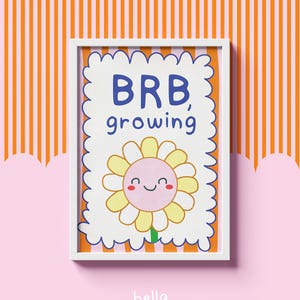 May include: A framed art print with a cheerful sunflower illustration and the text "BRB, growing" in blue. The background is a white scalloped shape with an orange border. The print is set against a pink and orange striped background.
