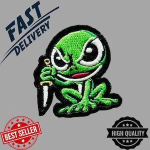 Puede incluir: Parche bordado con una rana verde con expresión enfadada, sosteniendo un cuchillo. La rana tiene ojos blancos y boca roja, delineada en negro. El parche está rodeado por un borde negro. El texto en la imagen dice "FAST DELIVERY", "BEST SELLER" y "HIGH QUALITY".