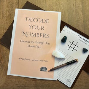 May include: A book titled "DECODE YOUR NUMBERS" with the subtitle "Discover the Energy That Shapes You." The book is accompanied by a notepad, pen, and crystals. The notepad features a tic-tac-toe grid and the words "Numerology Chart."