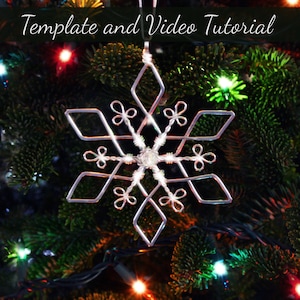 May include: A silver wire snowflake ornament hanging on a Christmas tree with green needles and colourful lights. The ornament is a simple design with six points and a small bead in the centre.