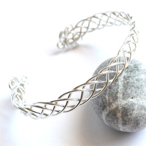 May include: A silver-colored cuff bracelet with a woven design. The open-ended bracelet is resting on a gray stone against a white background. The woven pattern is intricate and detailed, creating a textured appearance.