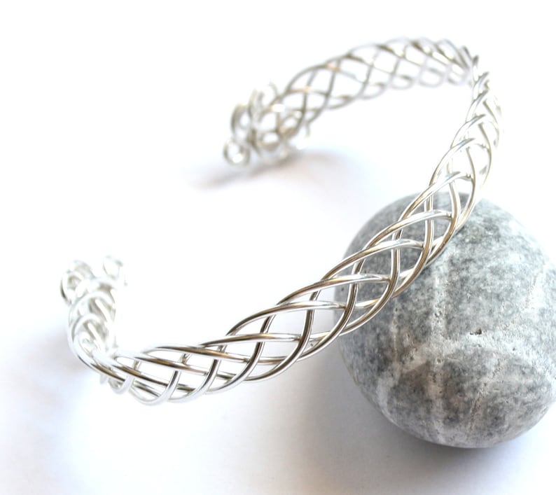 May include: A silver-colored cuff bracelet with a woven design. The open-ended bracelet is resting on a gray stone against a white background. The woven pattern is intricate and detailed, creating a textured appearance.