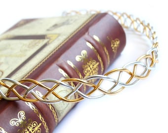 Puede incluir: Una corona trenzada de alambre dorado y plateado que descansa sobre un libro cerrado, encuadernado en cuero marrón con detalles dorados.