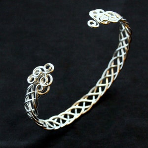 May include: A silver-colored cuff bracelet with a Celtic knot design. The bracelet features intricate woven patterns and decorative swirls at each end. The jewelry is set against a black background.