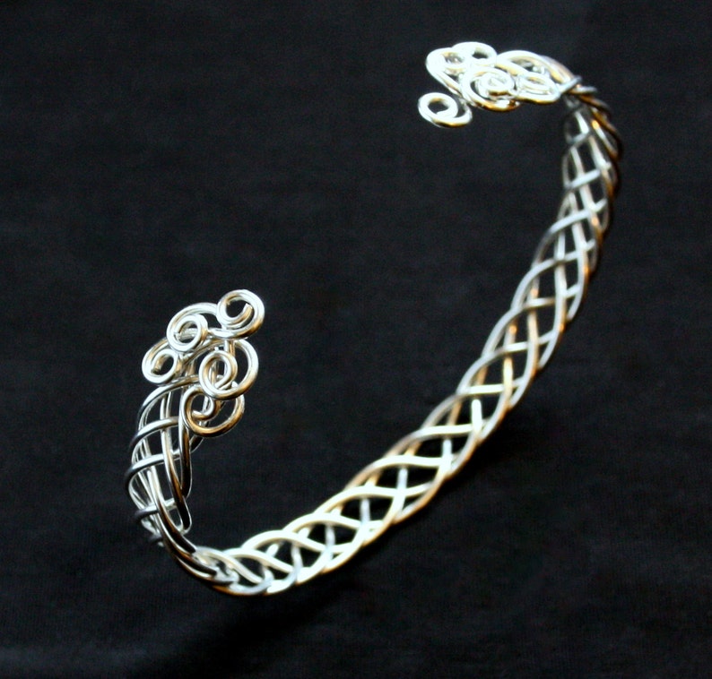 May include: A silver-colored cuff bracelet with a Celtic knot design. The bracelet features intricate woven patterns and decorative swirls at each end. The jewelry is set against a black background.