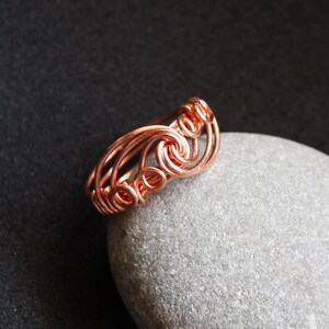 May include: A copper wire ring with a swirling design. The ring is on a gray stone background.