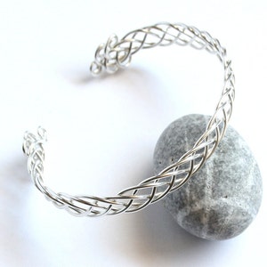 May include: A silver-colored cuff bracelet with a woven design. The bracelet is open at one end and rests on a gray stone against a white background. The woven pattern is intricate and detailed.
