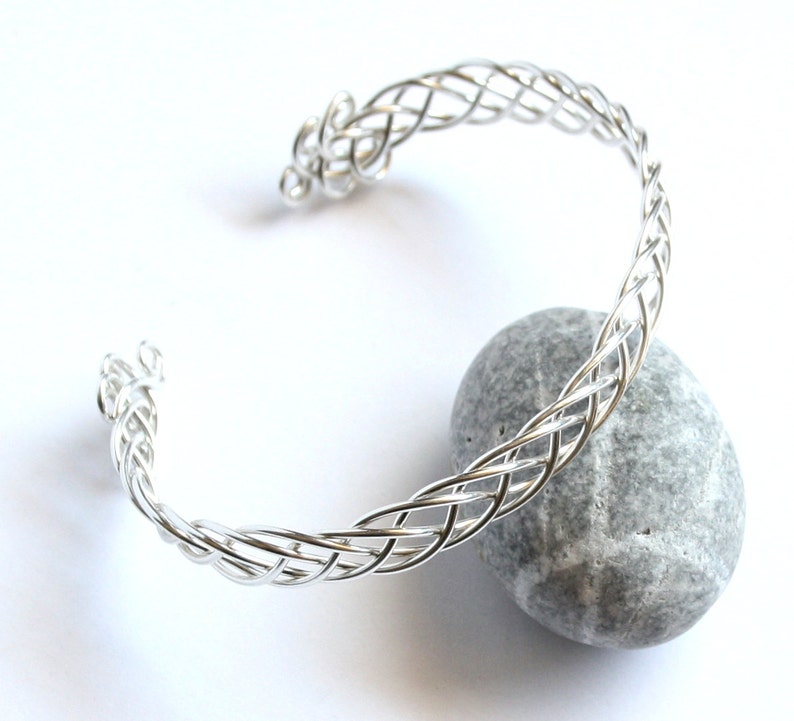 May include: A silver-colored cuff bracelet with a woven design. The bracelet is open at one end and rests on a gray stone against a white background. The woven pattern is intricate and detailed.