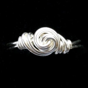 Könnte beinhalten: Ein silberfarbener Drahtring mit einem Spiraldesign. Der Ring hat eine zentrale Wirbel und gewickelte Drahtdetails, vor einem schwarzen Hintergrund. Der Ring ist ein Schmuckstück.