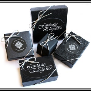 May include: An assortment of black jewelry boxes tied with silver ribbon. The boxes are various sizes and shapes, some with a textured pattern. The boxes have the text "Fantasia Elegance Artisan Jewelry" and "Handcrafted in Virginia" on them.