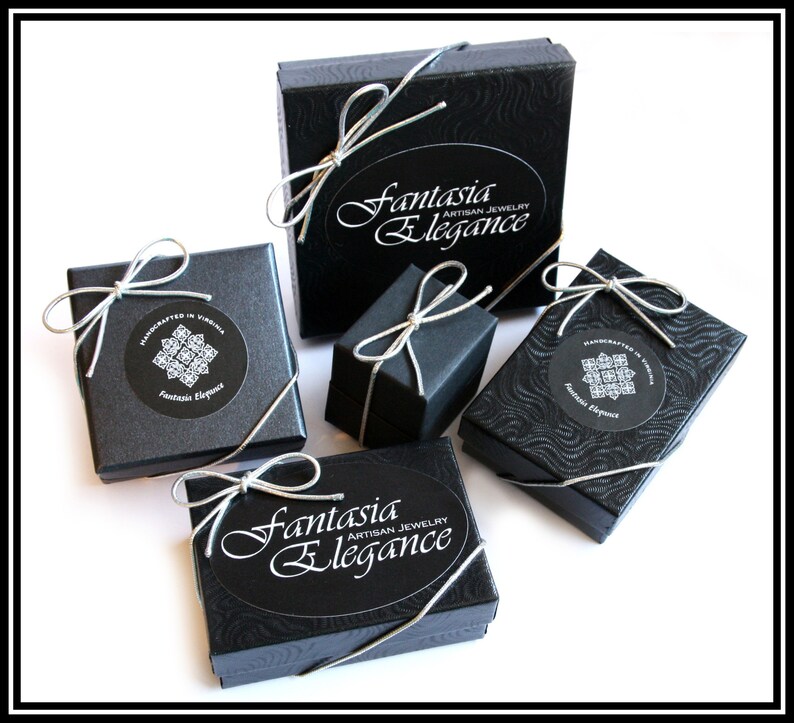 May include: An assortment of black jewelry boxes tied with silver ribbon. The boxes are various sizes and shapes, some with a textured pattern. The boxes have the text "Fantasia Elegance Artisan Jewelry" and "Handcrafted in Virginia" on them.