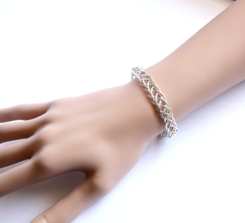 May include: A silver-colored woven bracelet is displayed on a person's wrist. The bracelet has a continuous, open weave design. The background is a bright white, highlighting the jewelry's intricate pattern and metallic sheen. The bracelet is a piece of jewelry.
