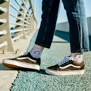 Puede incluir: Zapatillas deportivas negras y blancas con cordones blancos y una franja marrón. Las zapatillas se combinan con calcetines grises claros con un diseño de arcoíris colorido. La persona lleva vaqueros azules.