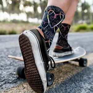 Puede incluir: Calcetines negros con un patrón de corazones coloridos se ven en los pies de una persona. La persona lleva zapatillas negras y monta en monopatín por una carretera pavimentada. Los calcetines, con el diseño de corazones, son el foco.