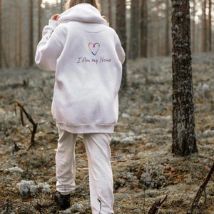 Puede incluir: Una persona con una sudadera con capucha blanca y un pantalón de chándal gris caminando por un bosque. La sudadera tiene un corazón arcoíris y el texto "I Am my Home". La persona lleva botas marrones oscuras.
