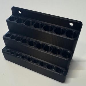 May include: A black, tiered tool organizer with multiple circular holes for storage. The organizer has three levels, each with a row of holes, and a top section with two mounting holes. The item is made of a solid, matte black material.